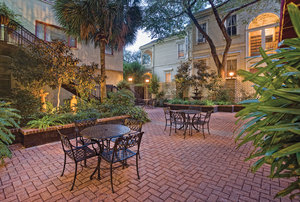 A brick patio area with metal tables and chairs, surrounded by trees and plants, with a building in the background.